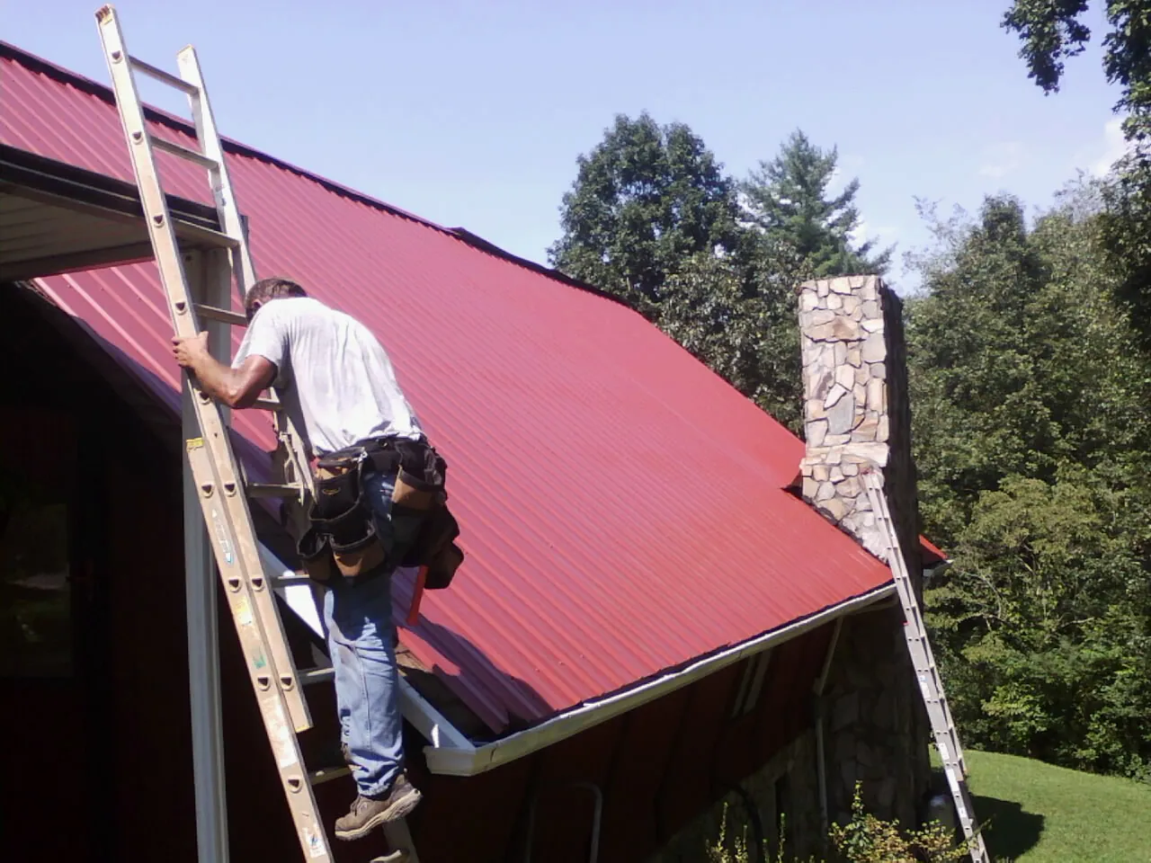 Roofer on a ladder beside a red metal roof and stone chimney — Soffit & Fascia Repair by Roofing Brothers in New Durham